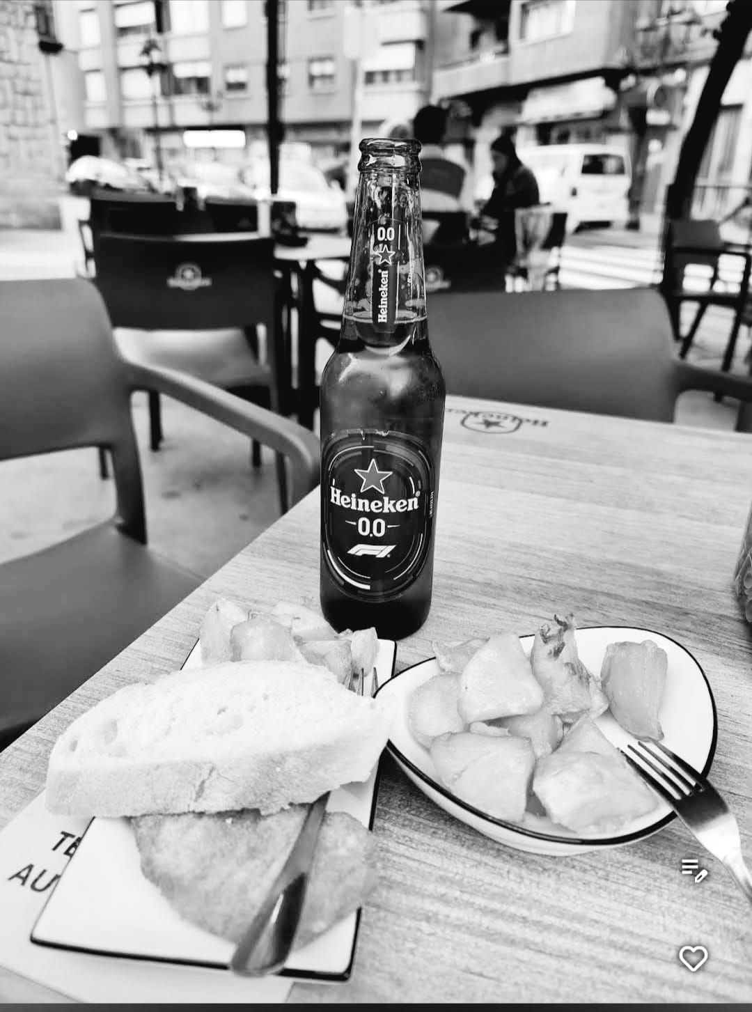 Fotografía en blanco y negro de una botella de cerveza, tapas y pan en la mesa de un restaurante al aire libre.
