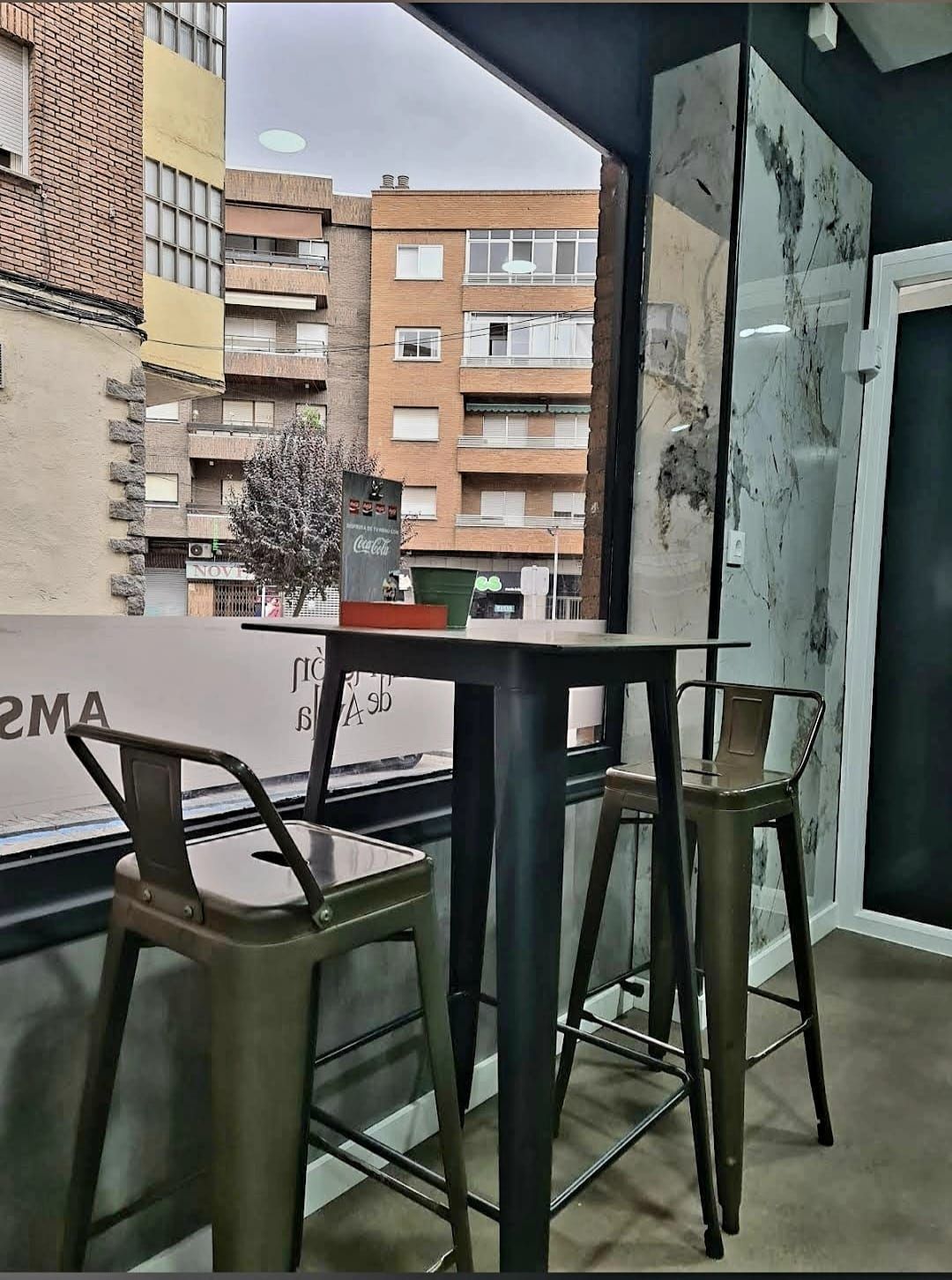 Interior de cafetería con mesa alta, dos taburetes, vista de edificios fuera de la ventana.
