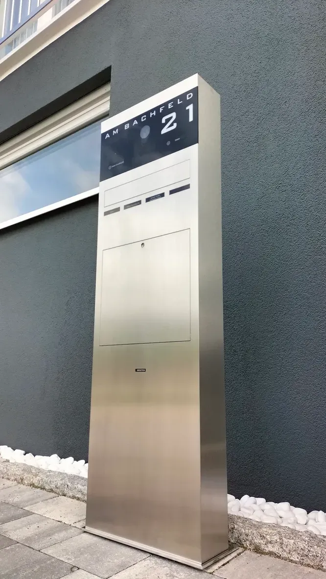 Vor einem Gebäude steht ein Briefkasten aus Edelstahl.
