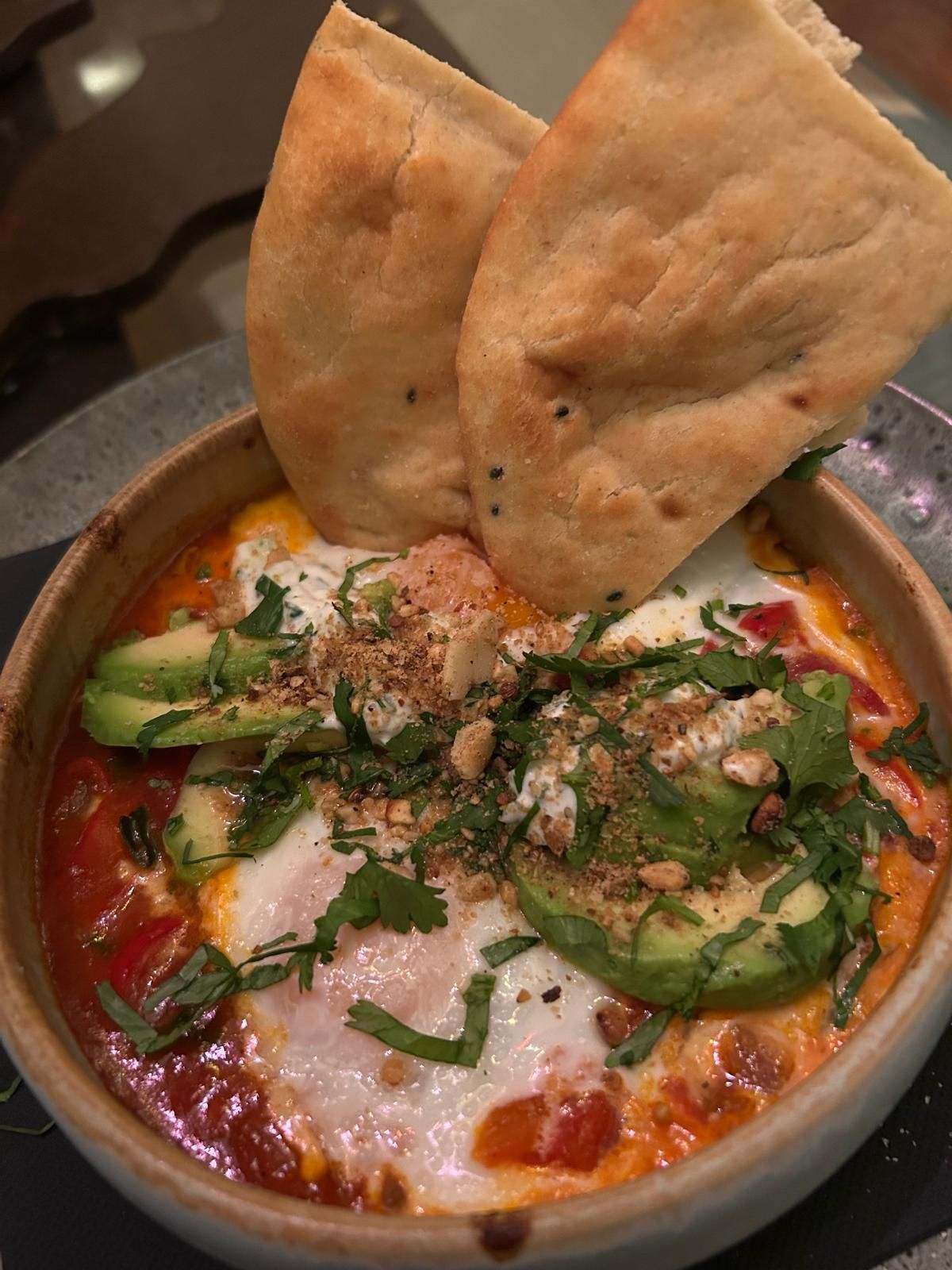 Shakshuka con aguacate, hierbas y pan plano en un tazón de madera.