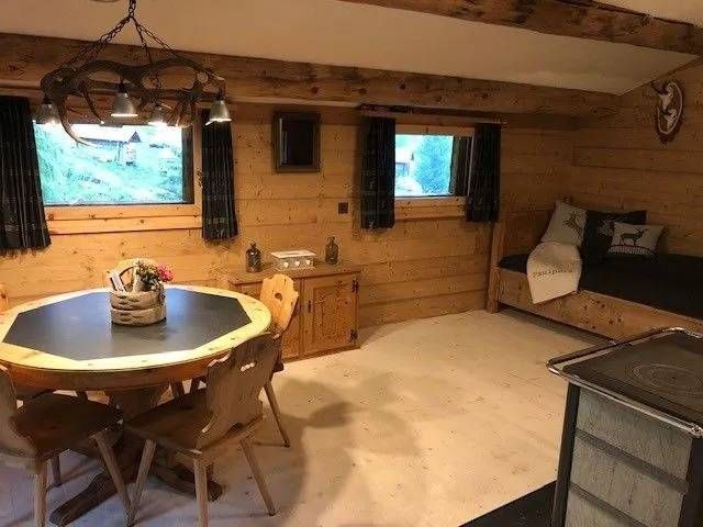 Gemütliches Hütteninterieur mit Holzwänden und -möbeln, rundem Tisch, kleinen Fenstern und einem Tagesbett.