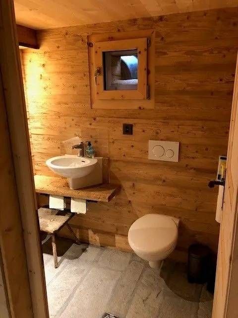 Holzbadezimmer mit Toilette, Waschbecken und kleinem Fenster. Steinboden und Holzwände.