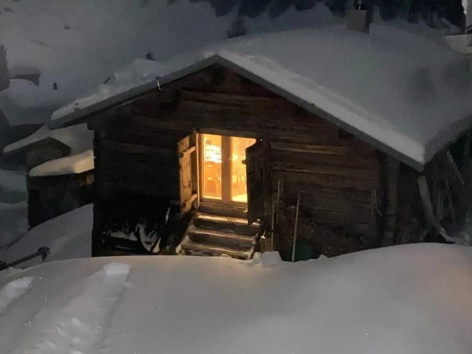 Eine schneebedeckte Holzhütte bei Nacht, durch deren offene Tür warmes Licht scheint.