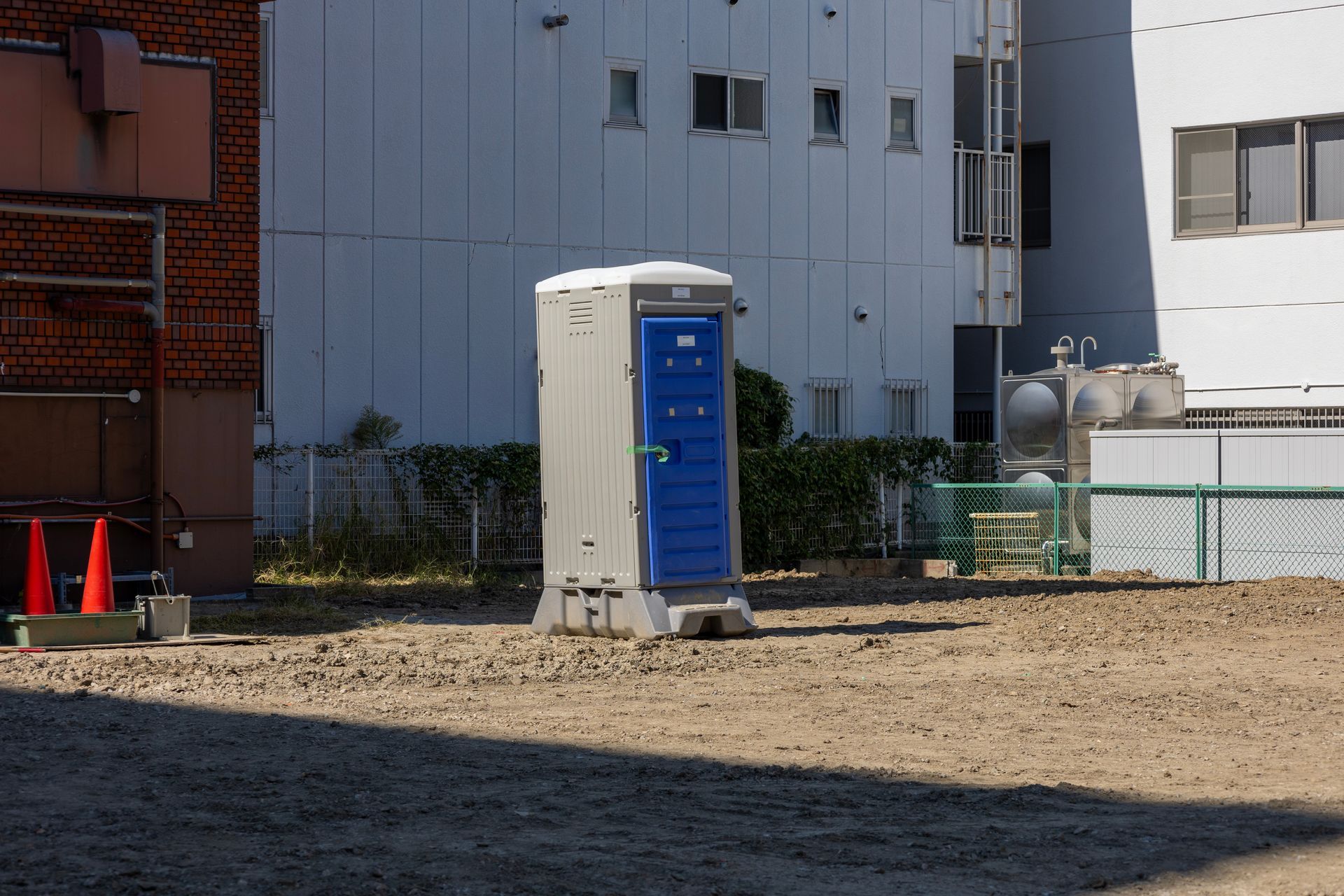 Toilettes portables bleues et grises sur du gravier, avec des bâtiments en arrière-plan.