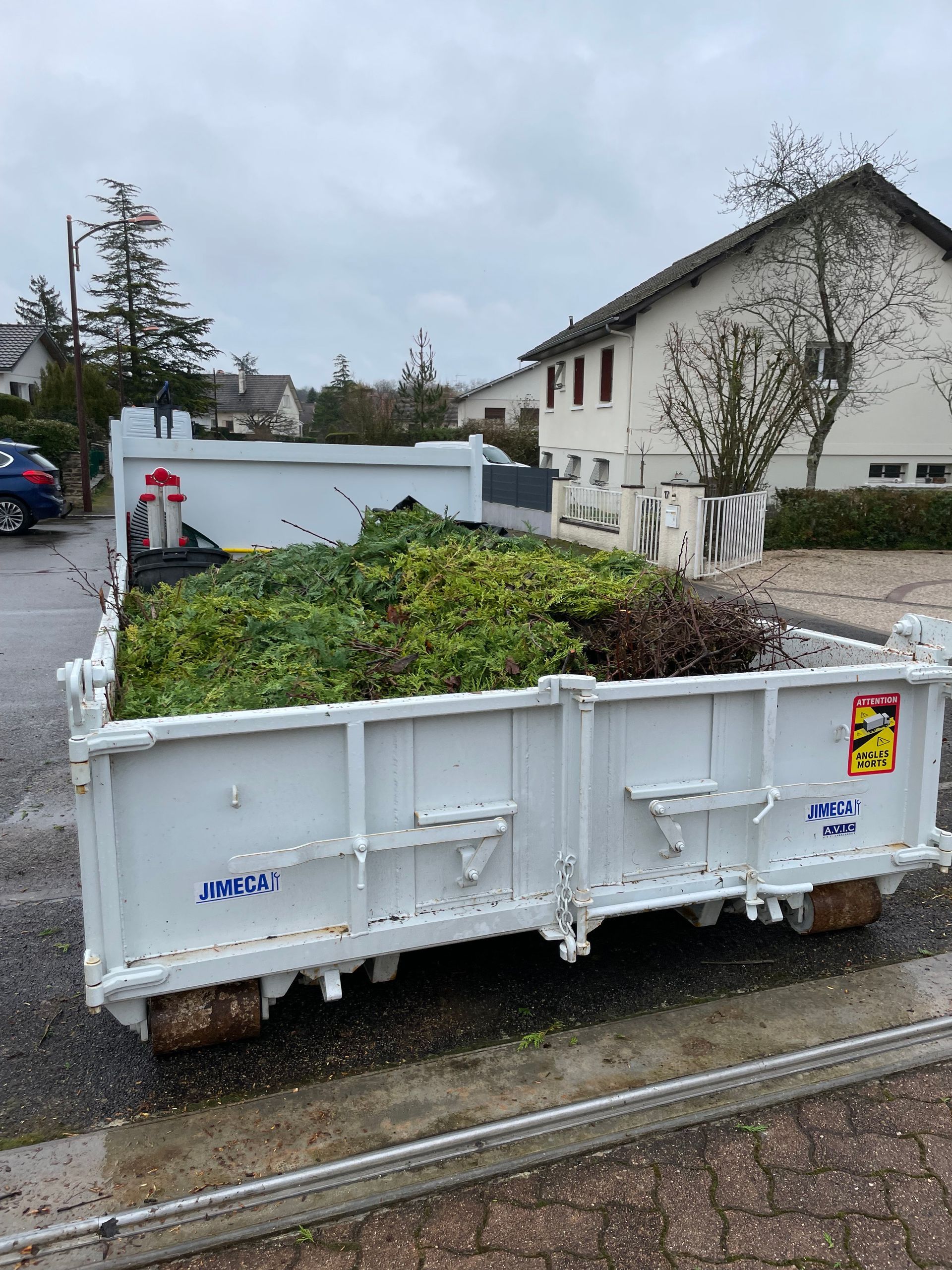 Un conteneur blanc rempli de déchets verts de jardin, stationné près d'une maison sous un ciel nuageux.