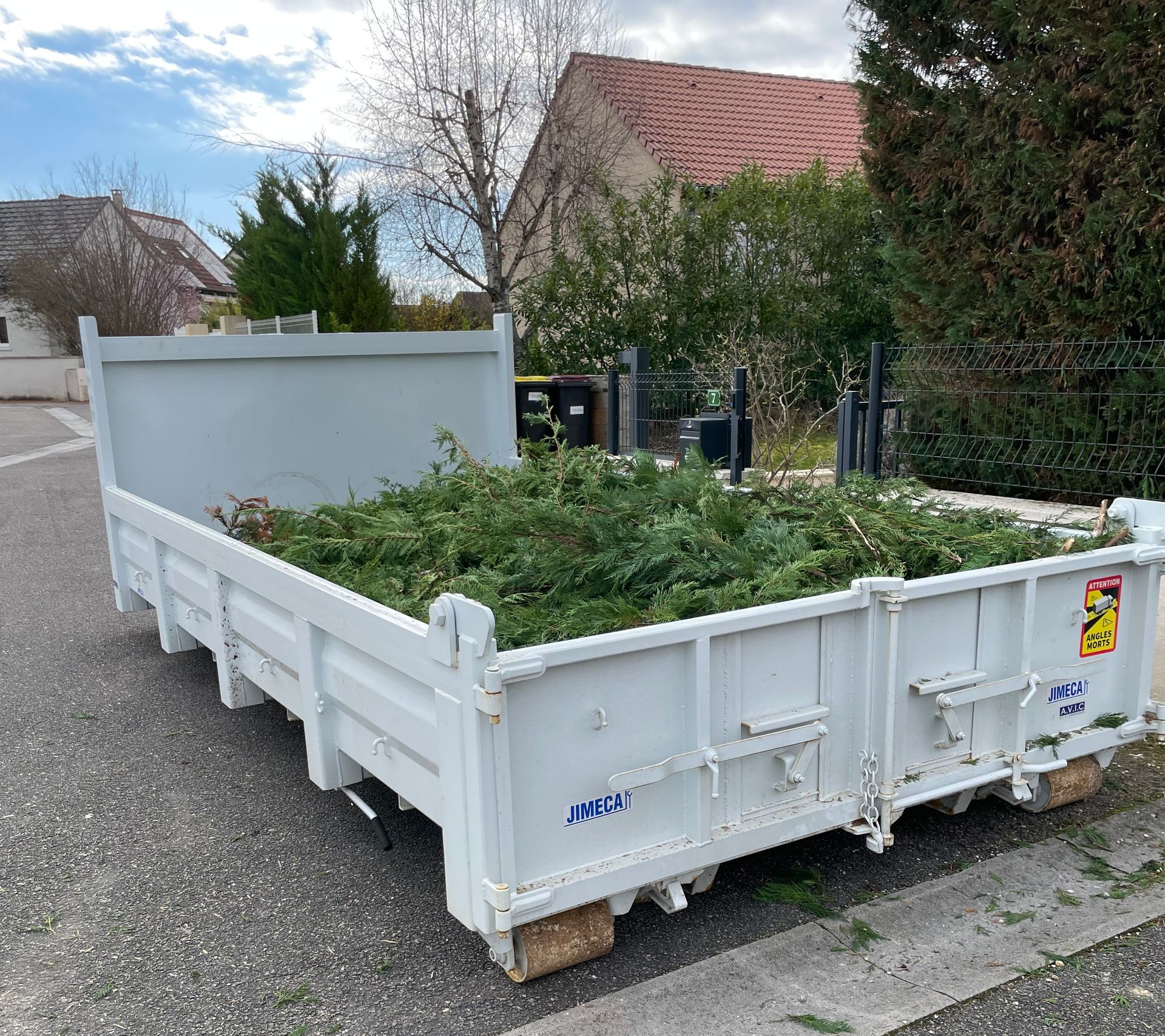 Un conteneur à déchets métalliques blancs rempli de déchets verts de jardin est stationné sur une route goudronnée.
