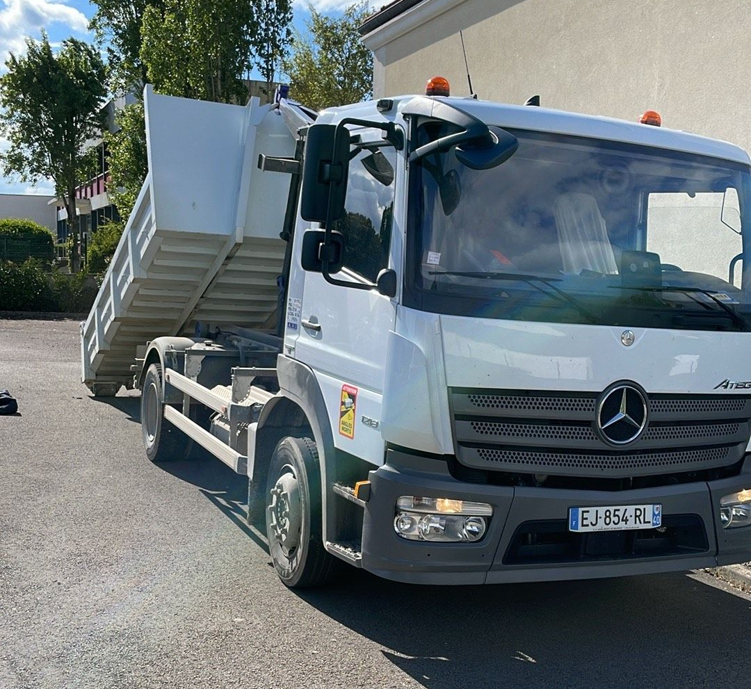 Camion-benne blanc à plateau surélevé, stationné sur une route goudronnée près d'un bâtiment.