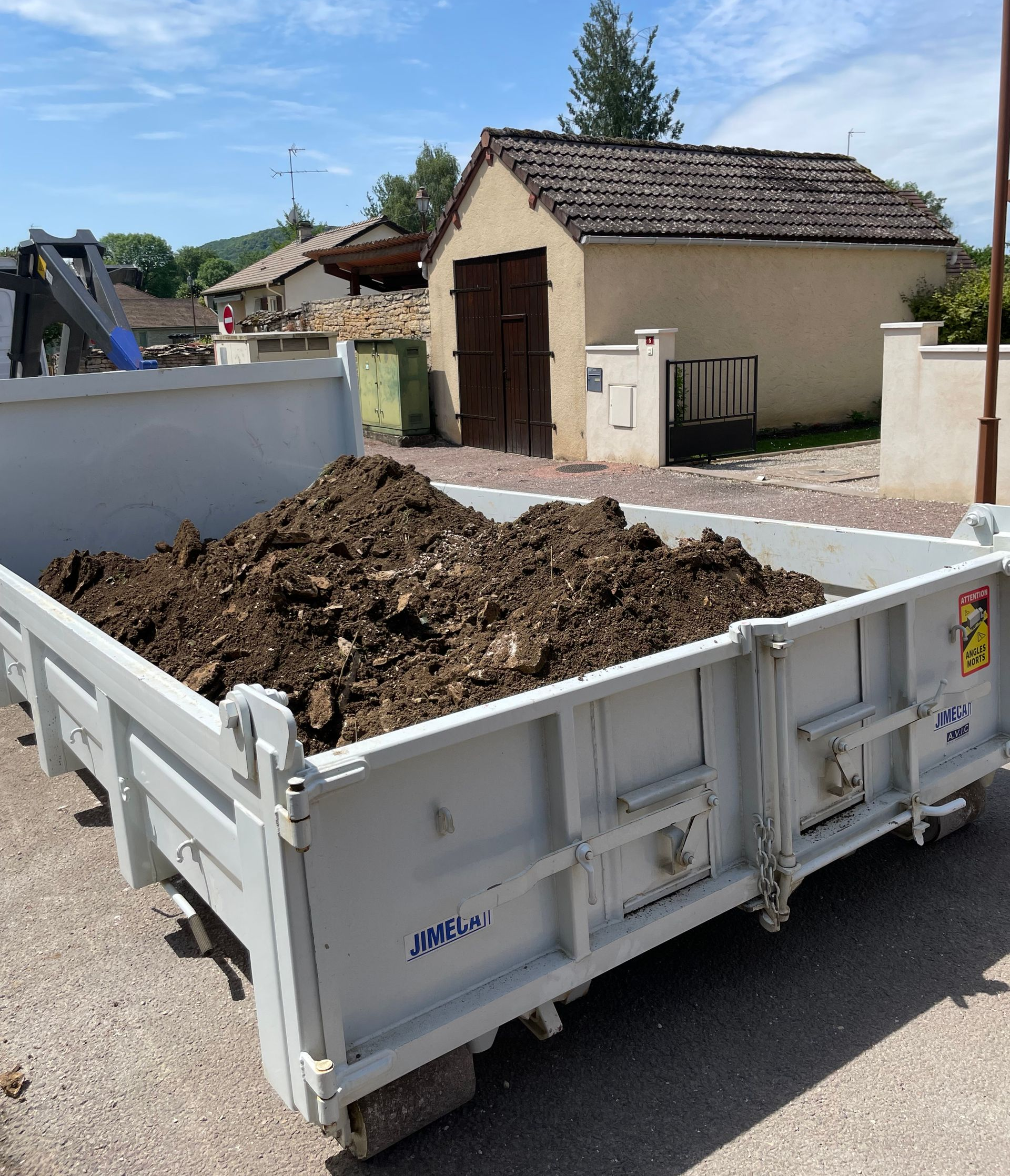 Une benne à ordures blanche remplie de terre sombre, placée à l'extérieur avec des bâtiments en arrière-plan.