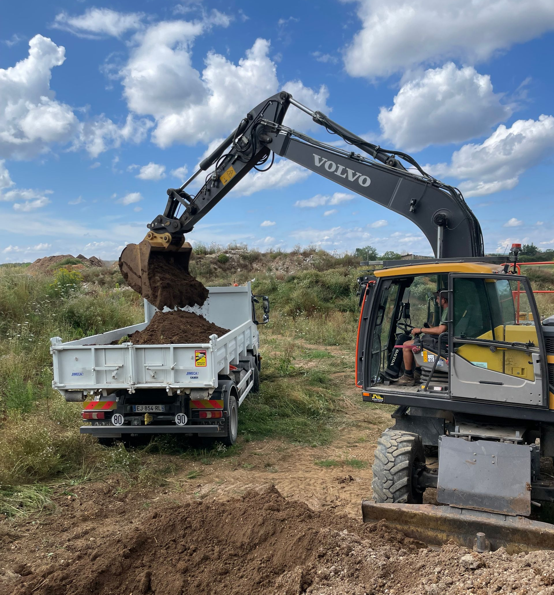 Une excavatrice charge de la terre dans la benne d'un camion blanc, dans un champ sous un ciel partiellement nuageux.