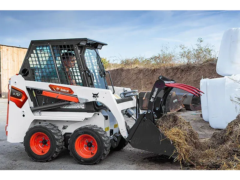 Chargeuse compacte Bobcat avec une personne à l'intérieur, ramassant du foin. Machine blanche et orange, à l'extérieur.