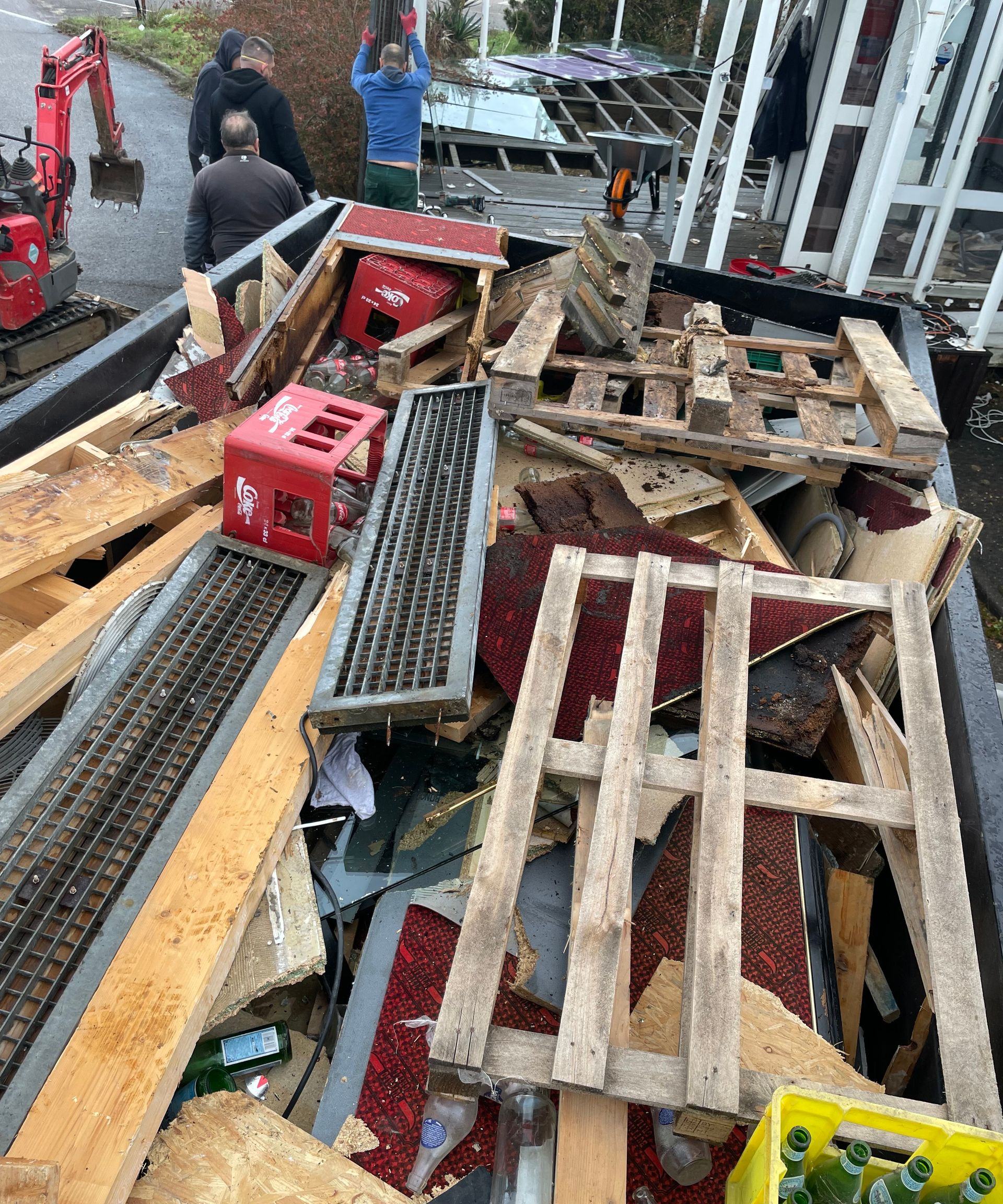 Des débris, dont du bois et des caisses rouges, se trouvent dans une remorque. Des personnes travaillent près d'une petite pelle mécanique et d'une structure partiellement démantelée.