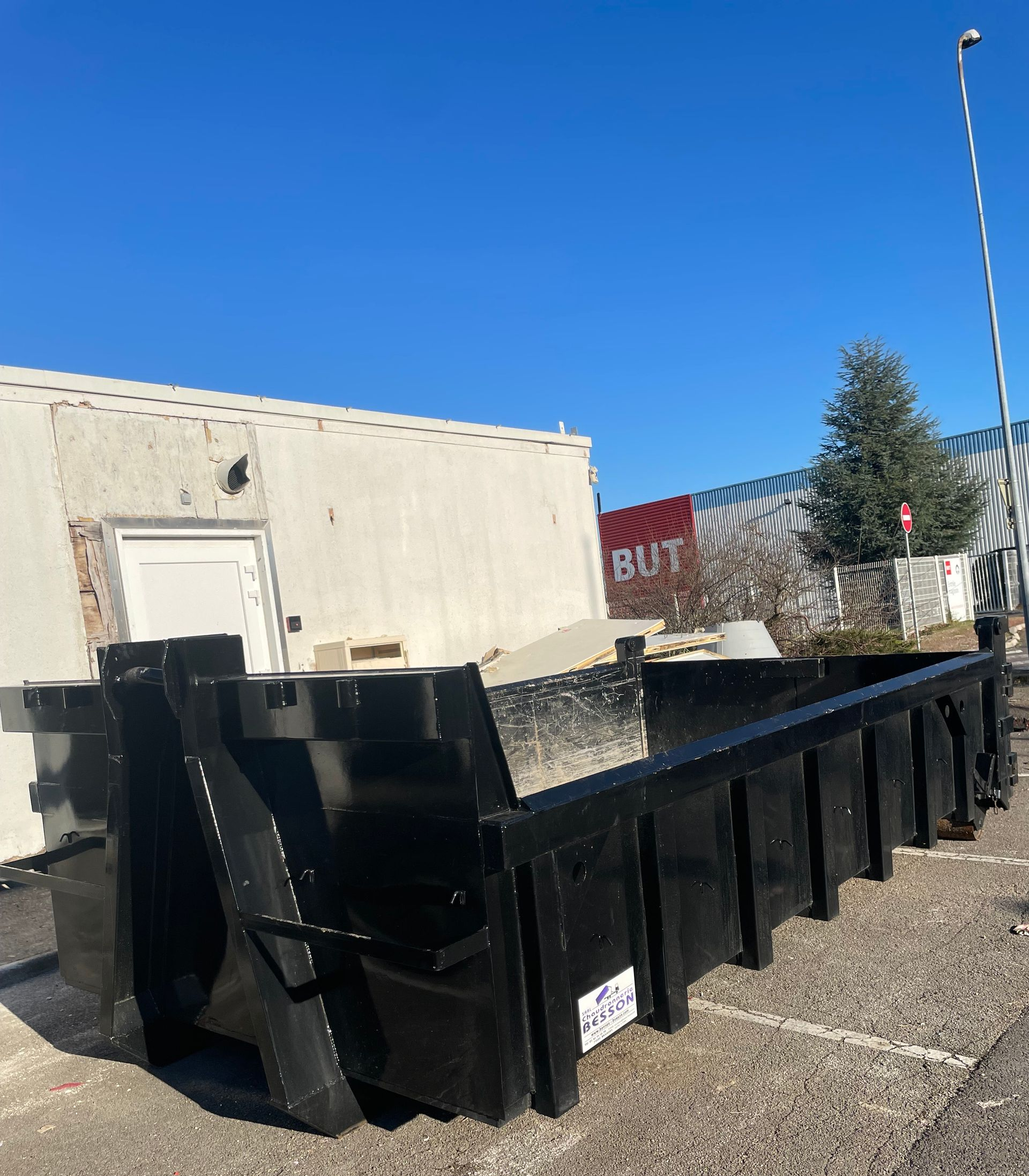 Une benne à ordures noire devant un bâtiment blanc sous un ciel bleu azur.
