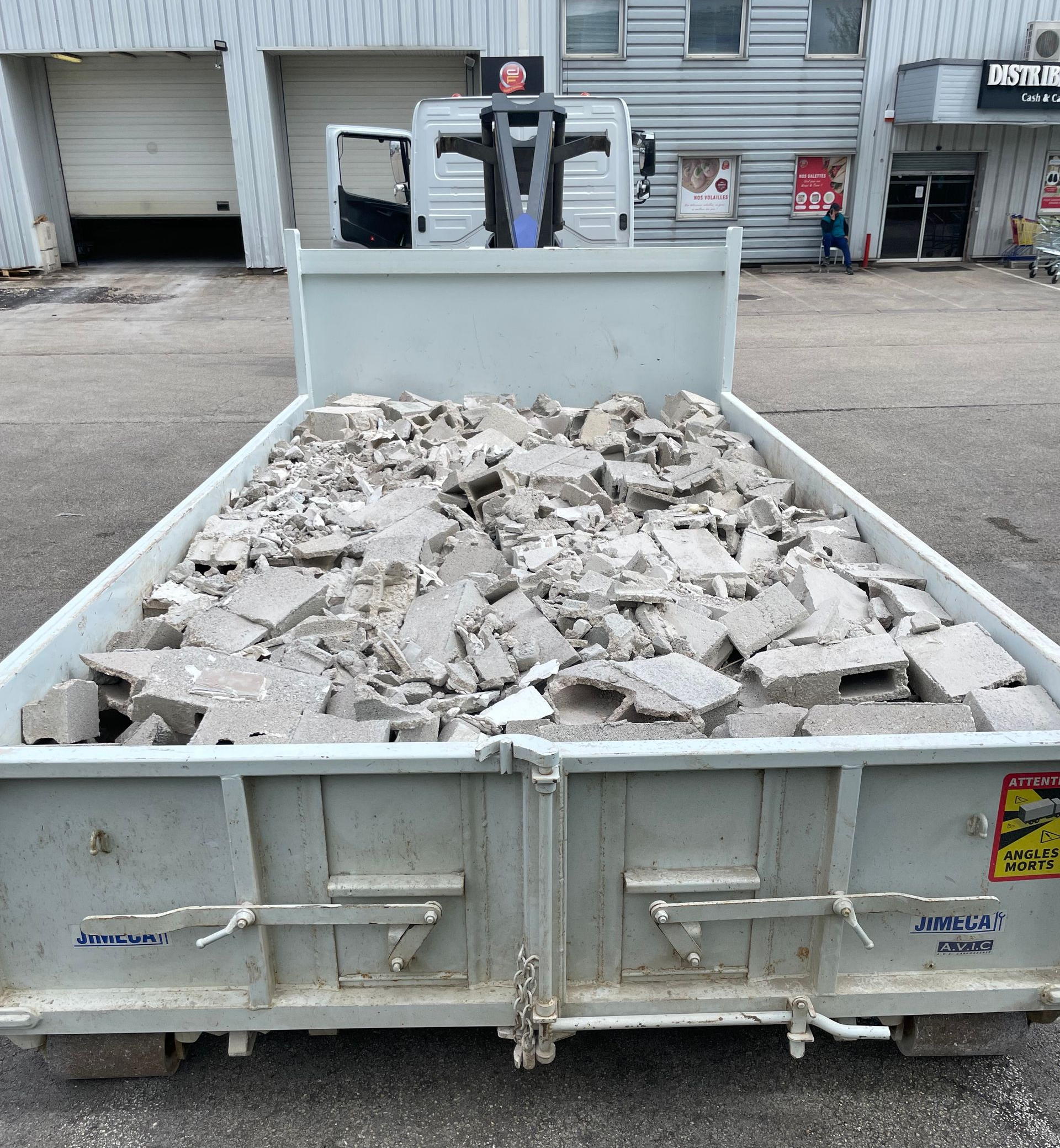 Une benne métallique remplie de débris de béton est stationnée devant un bâtiment.