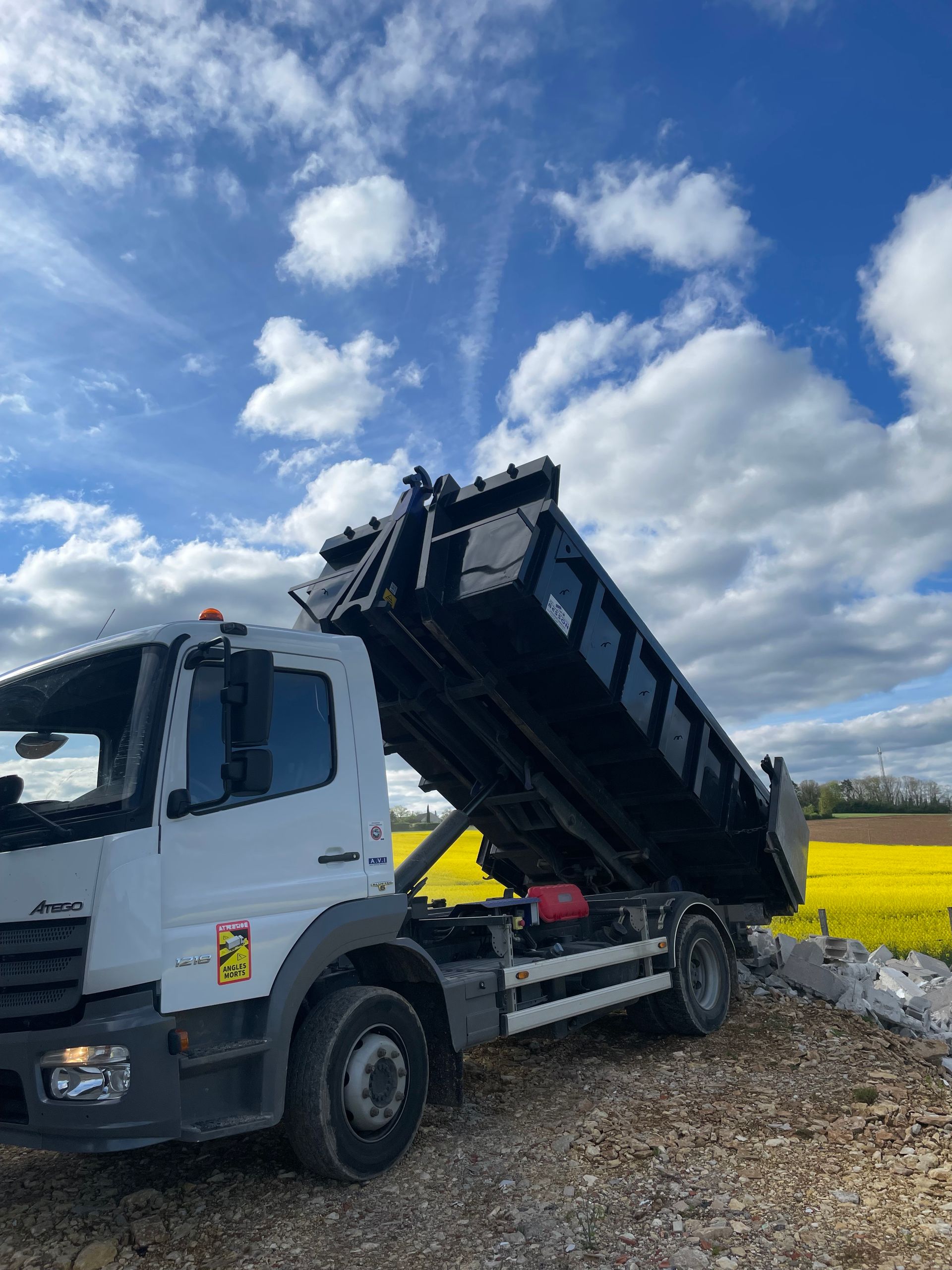 Camion-benne blanc à plateau noir surélevé, sol de gravier, champ jaune, ciel bleu.