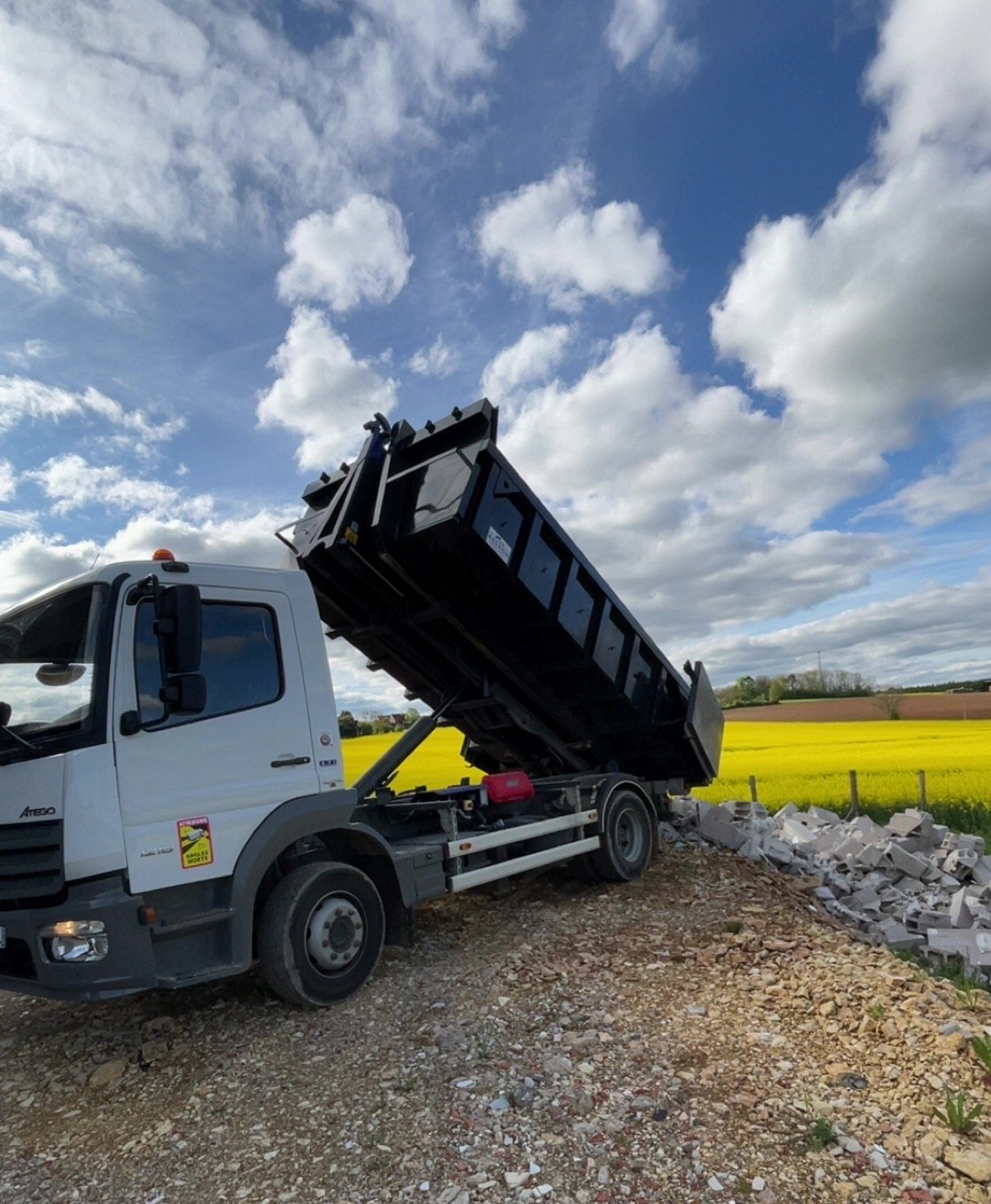 Camion-benne blanc à plateau noir surélevé, déversant des débris sur un chemin de gravier, ciel bleu avec des nuages en arrière-plan.
