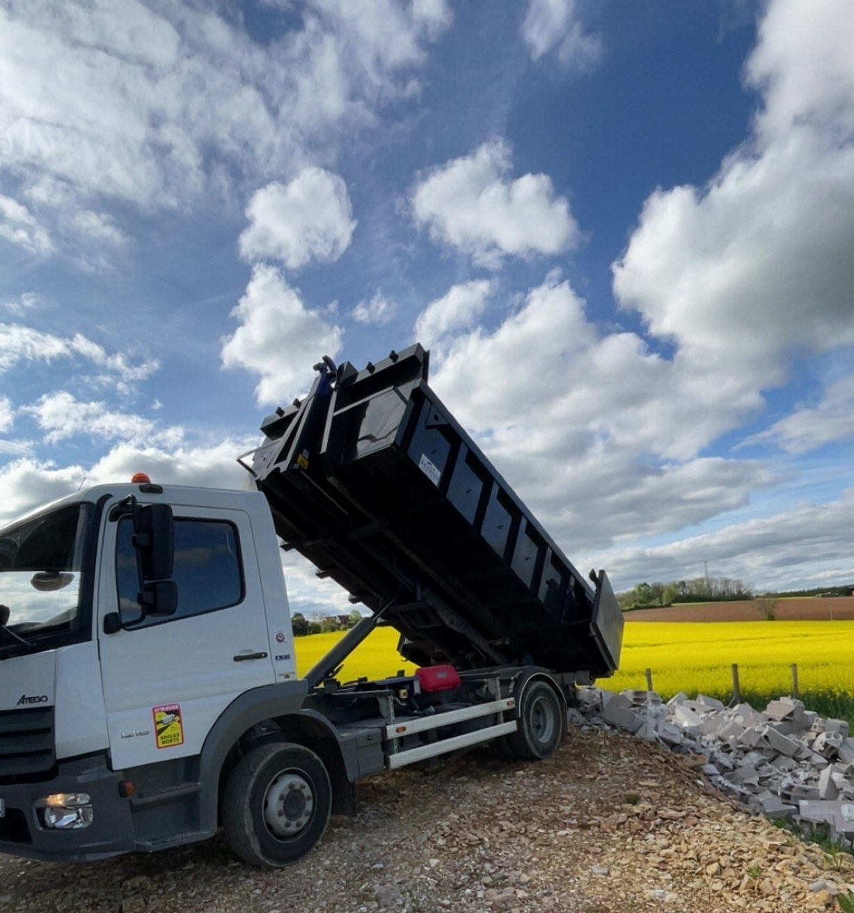 Camion-benne blanc à benne noire déversant des pierres sur un chantier ; champ jaune et ciel bleu en arrière-plan.