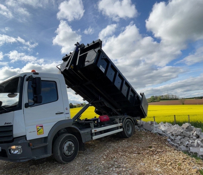 Un camion-benne blanc à plateau noir surélevé déverse des débris près d'un champ jaune sous un ciel nuageux.