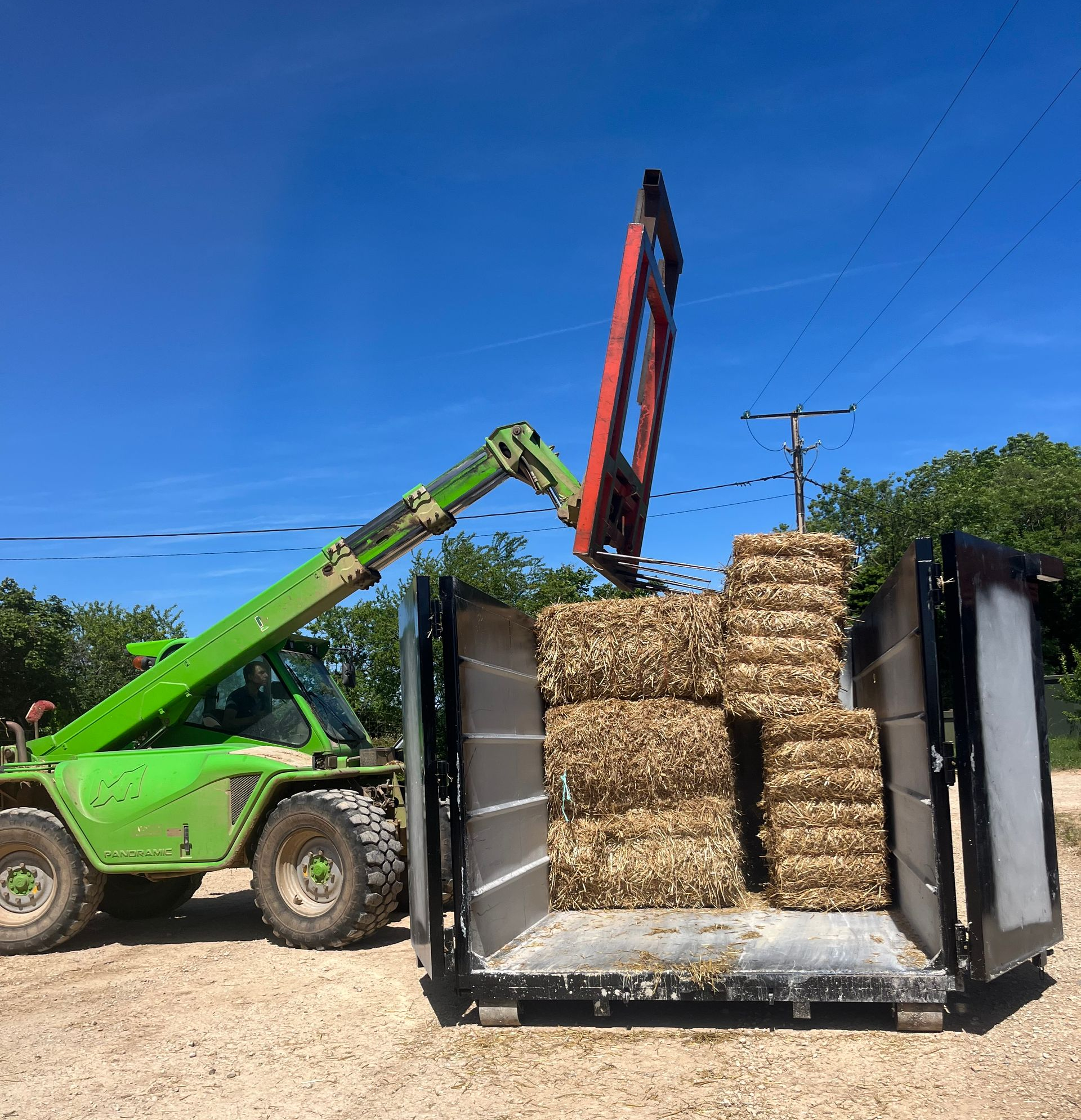 Un chariot télescopique vert charge des balles de foin dans une remorque par une journée ensoleillée.