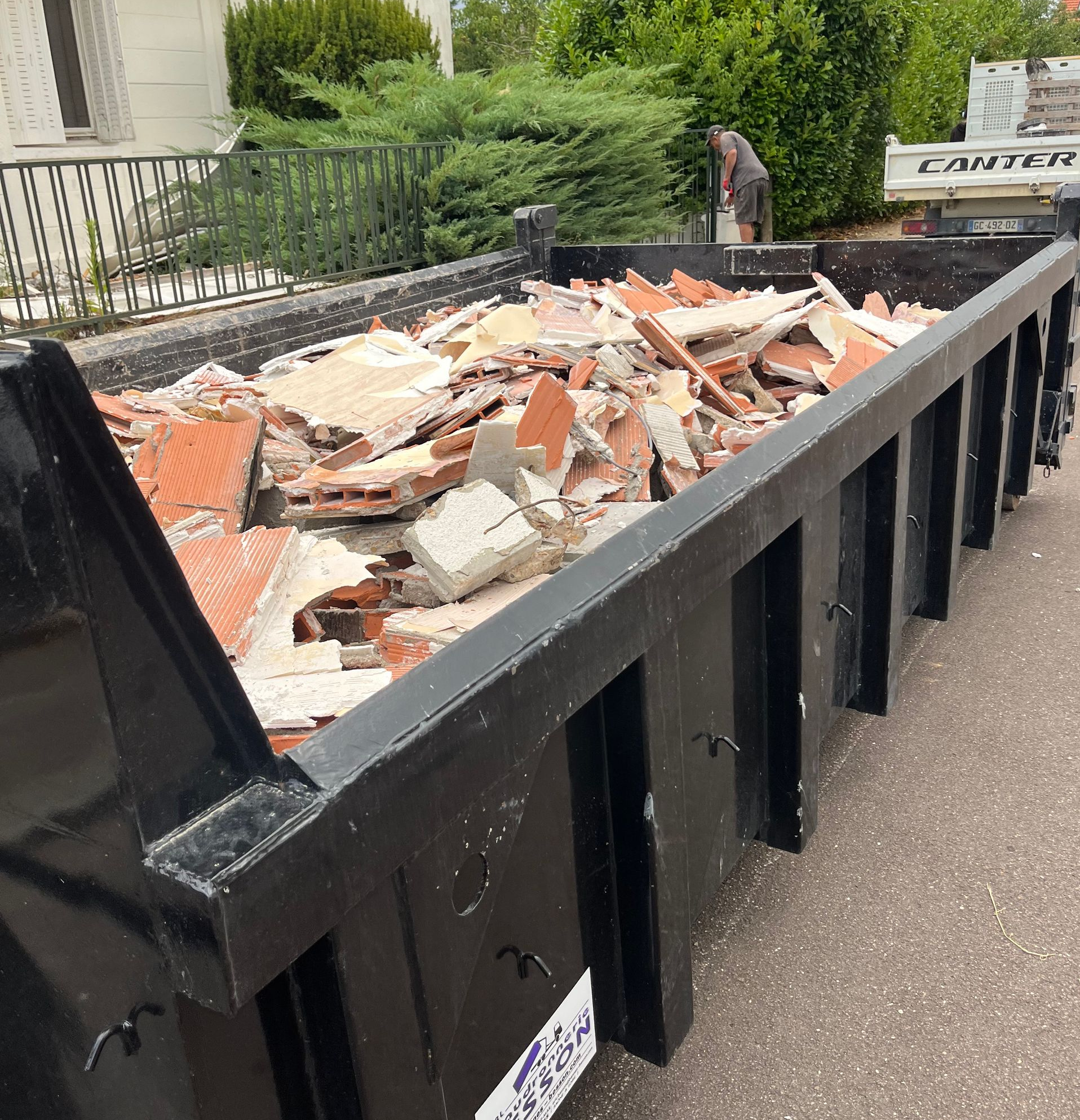 Benne noire remplie de débris de construction, une personne y charge d'autres gravats. Extérieur.