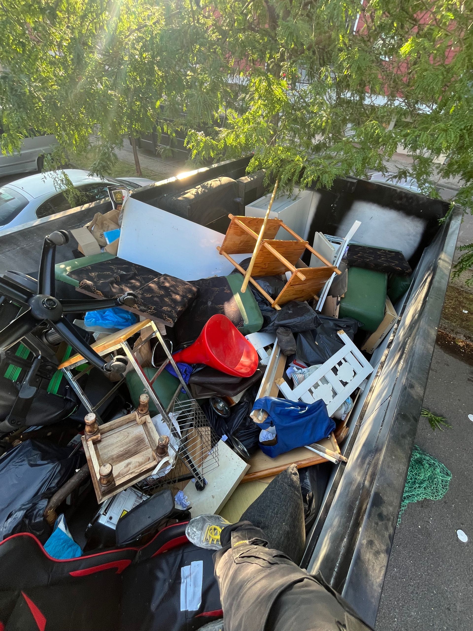 La benne à ordures déborde de divers objets jetés, notamment des meubles, du plastique et des déchets.