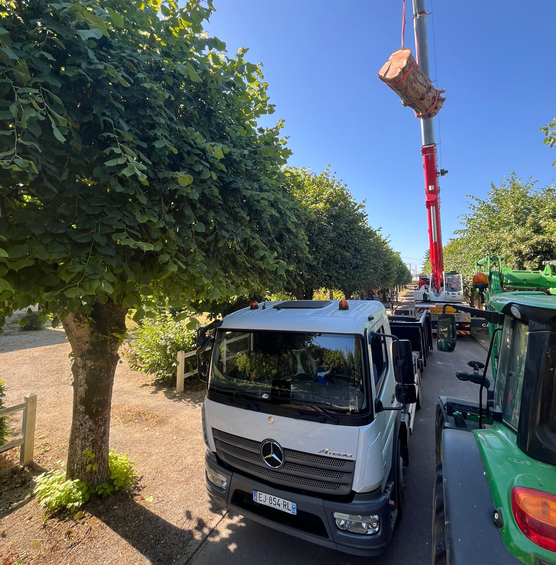 Un camion et un tracteur avec une grue soulèvent un tronc d'arbre dans une rue ensoleillée.