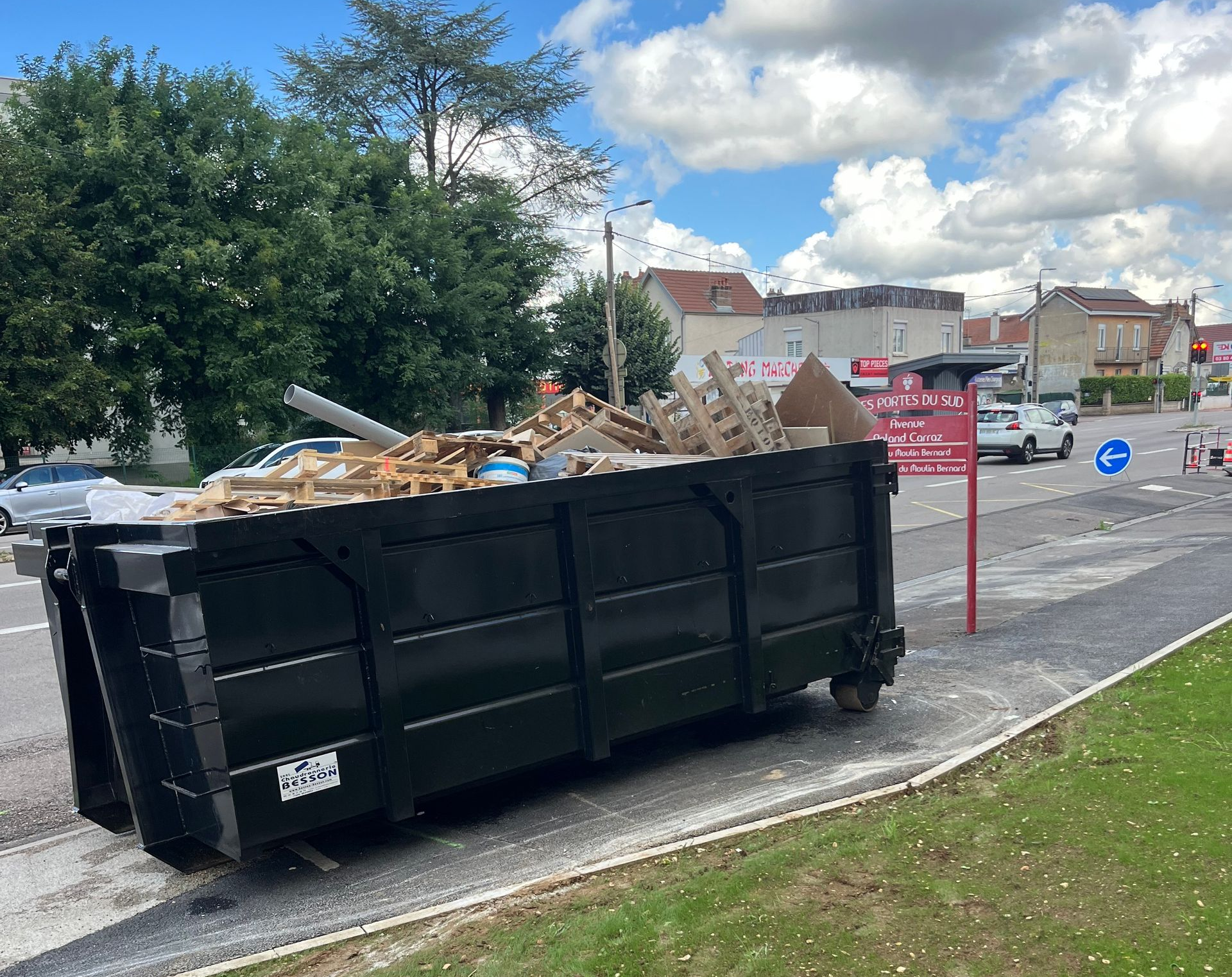 Une benne à ordures noire, débordant de débris de construction, se trouve sur un trottoir, au bord d'une route.