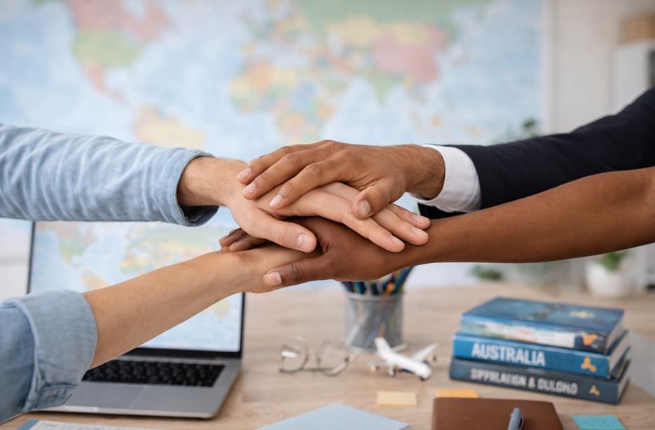Varias manos apiladas en un gesto de colaboración sobre un escritorio con una computadora portátil y libros de viajes.