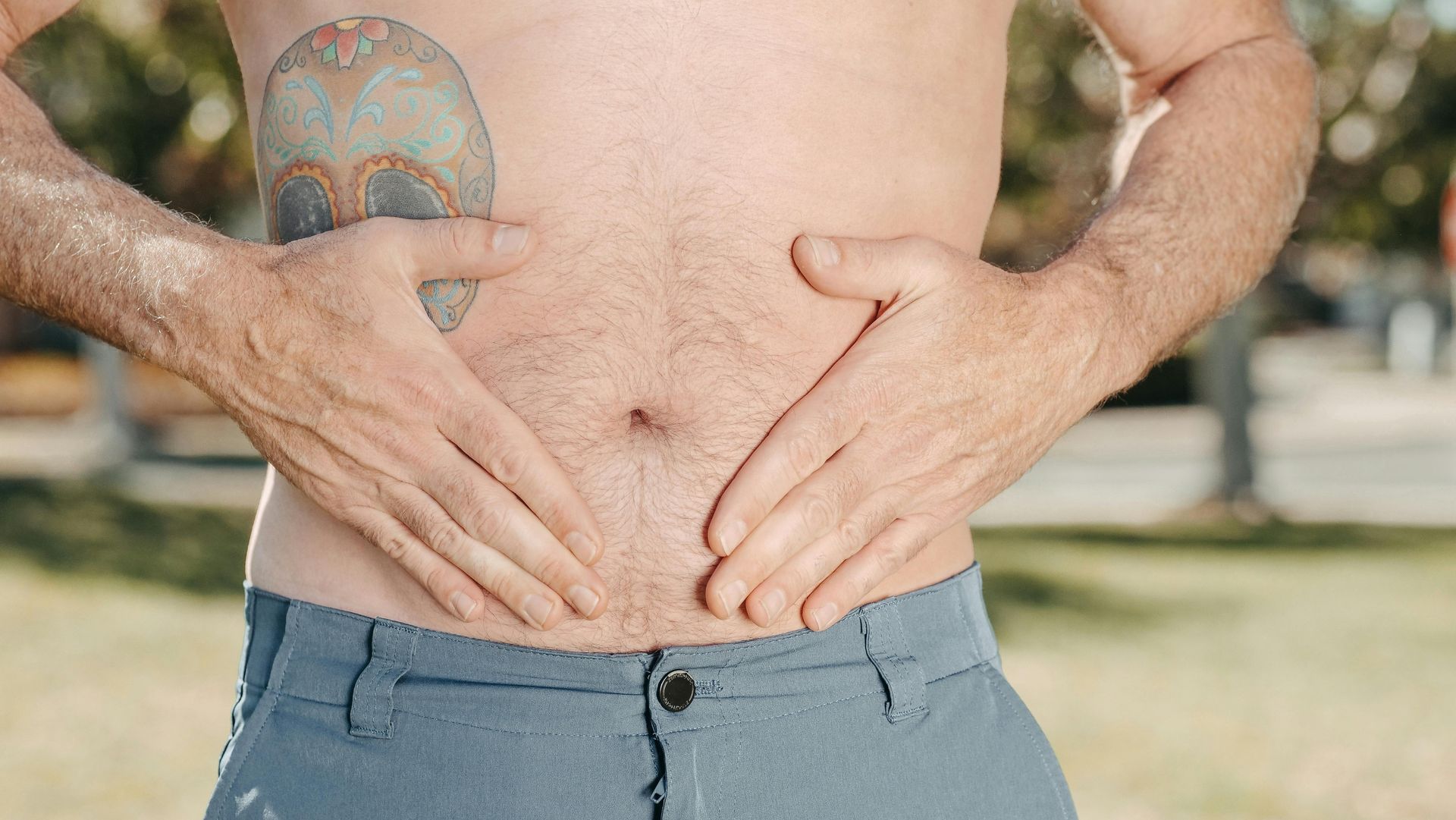 Ein Mann ohne Hemd und mit einer Tätowierung auf der Brust hält seinen Bauch mit den Händen.