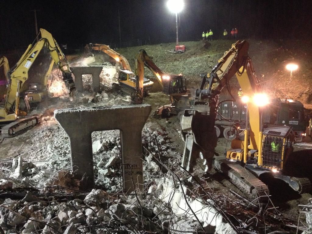 Chantier de construction la nuit, avec des excavatrices démolissant des piliers en béton sous des projecteurs puissants.