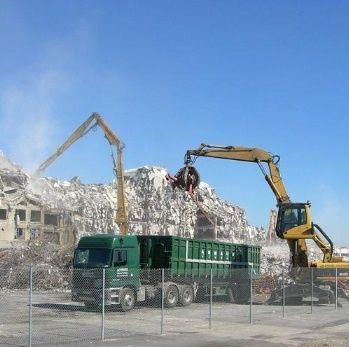 Chantier de démolition avec deux excavatrices et un camion ramassant les débris, sur fond de ciel bleu dégagé.