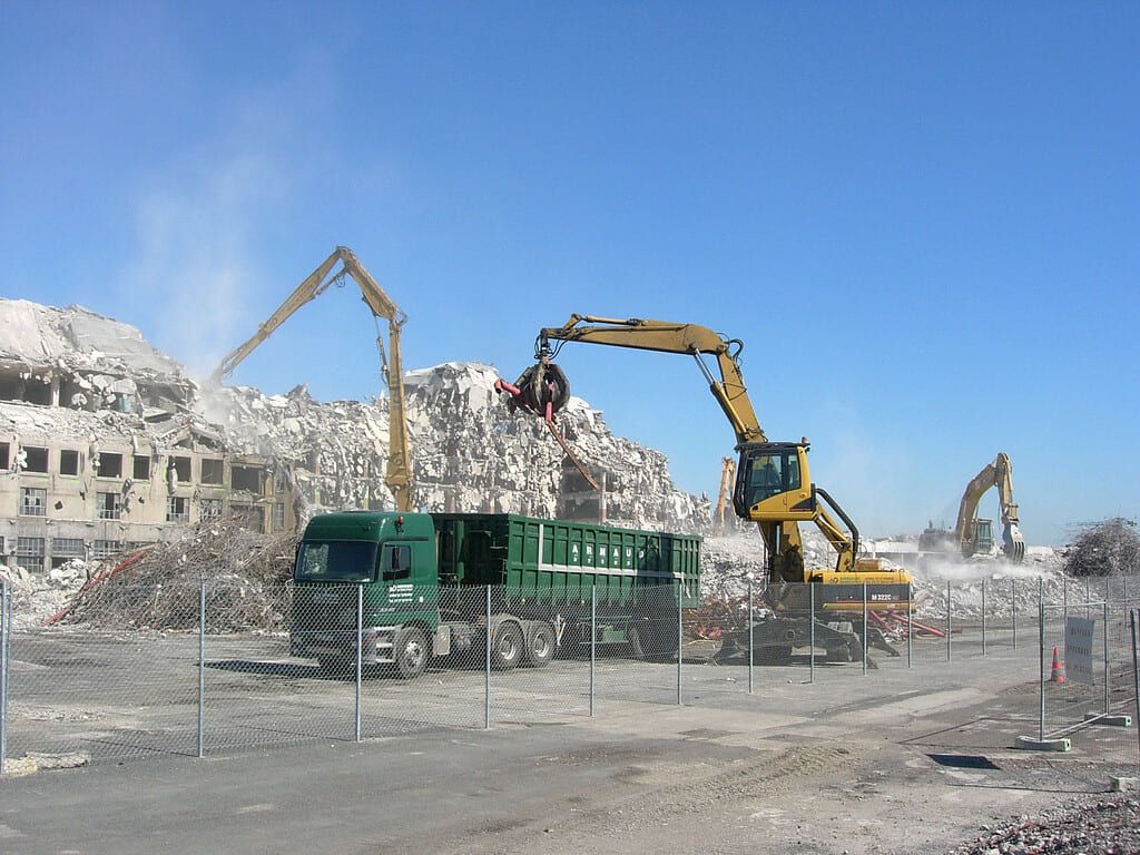 Chantier de construction avec des excavatrices en train de démanteler un bâtiment ; tas de débris et un camion-benne vert.