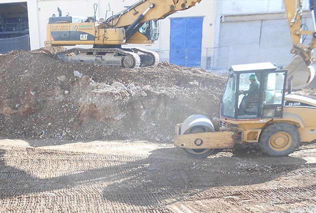Des engins de chantier jaunes compactent la terre près d'un grand tas de terre, avec une excavatrice en arrière-plan.