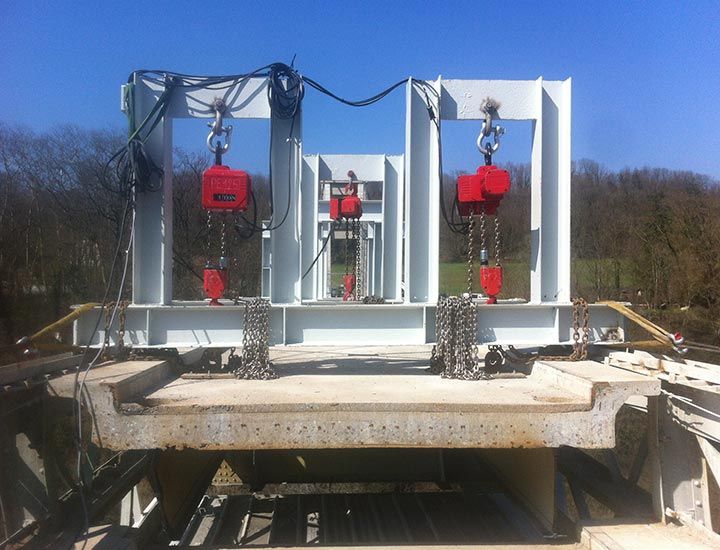 Une structure équipée de trois palans à chaîne rouges soulève une poutre en béton à l'extérieur, par une journée ensoleillée.