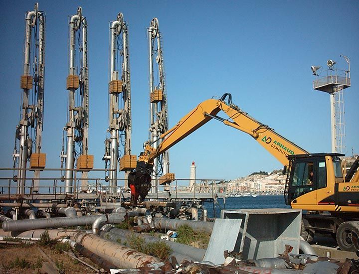 Une excavatrice sur un site industriel, à proximité de hauts pipelines et de structures, sous un ciel bleu dégagé.