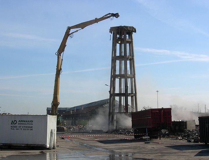 Une haute tour en béton est démolie par un bras hydraulique par une belle journée ensoleillée. On aperçoit des débris et de la poussière.