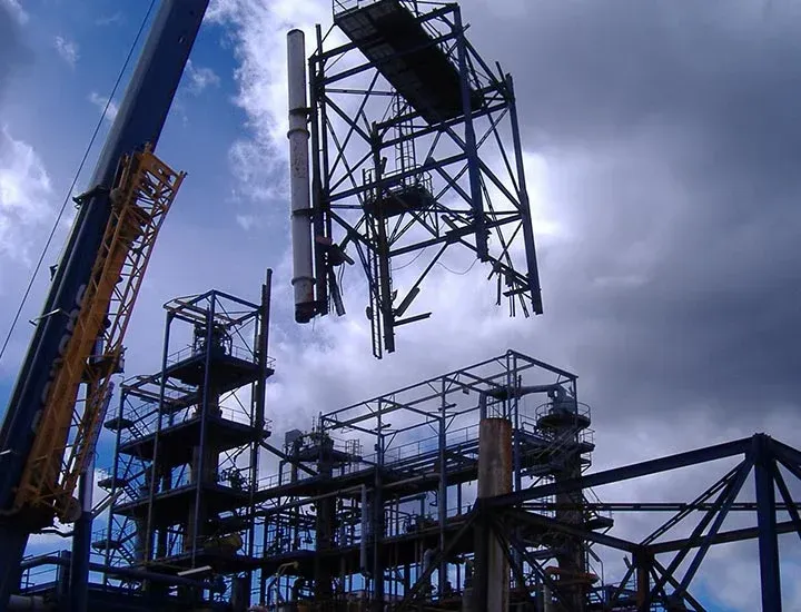 Grue soulevant une structure en acier sur un site industriel, ciel nuageux.