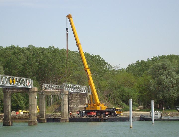 Une grue jaune soulève une section de pont métallique au-dessus de l'eau. Des arbres verts se dessinent à l'arrière-plan.