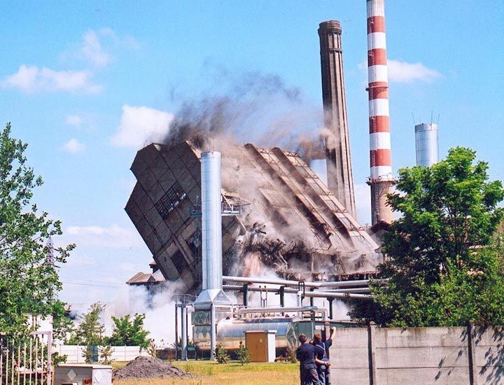 Un bâtiment d'usine s'effondre, dégageant de la fumée ; des cheminées se dressent à proximité. Ciel bleu.