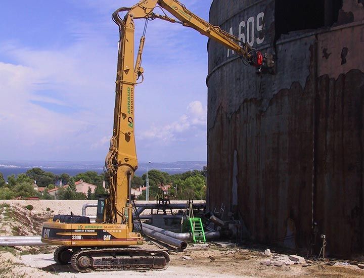 Une pelleteuse jaune démolit un grand réservoir métallique près de l'eau, du soleil et d'autres bâtiments.