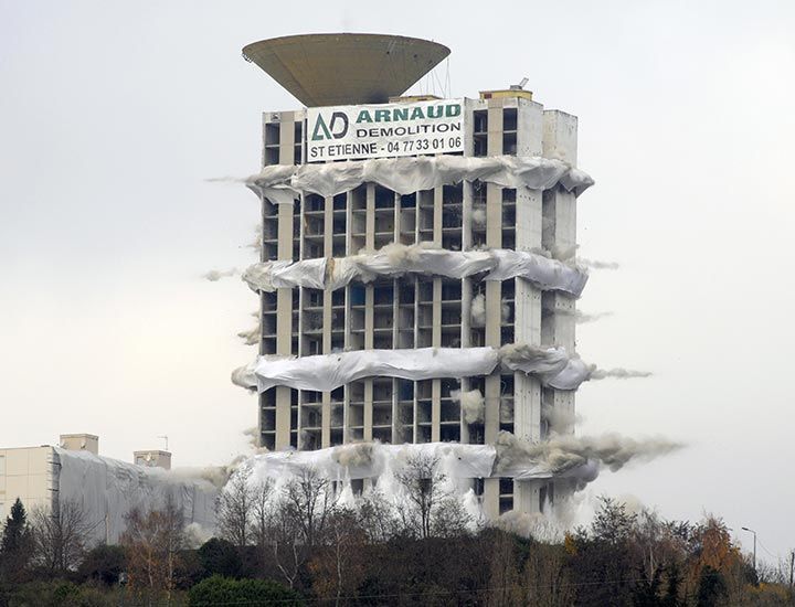 Implosion d'un bâtiment, partiellement recouvert de blanc, nuages ​​de poussière qui s'élèvent, ciel gris.