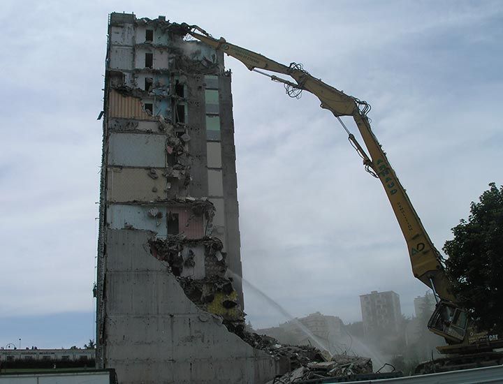 Un immeuble de grande hauteur est démoli par le bras d'une grande excavatrice jaune ; des débris de béton sont visibles.