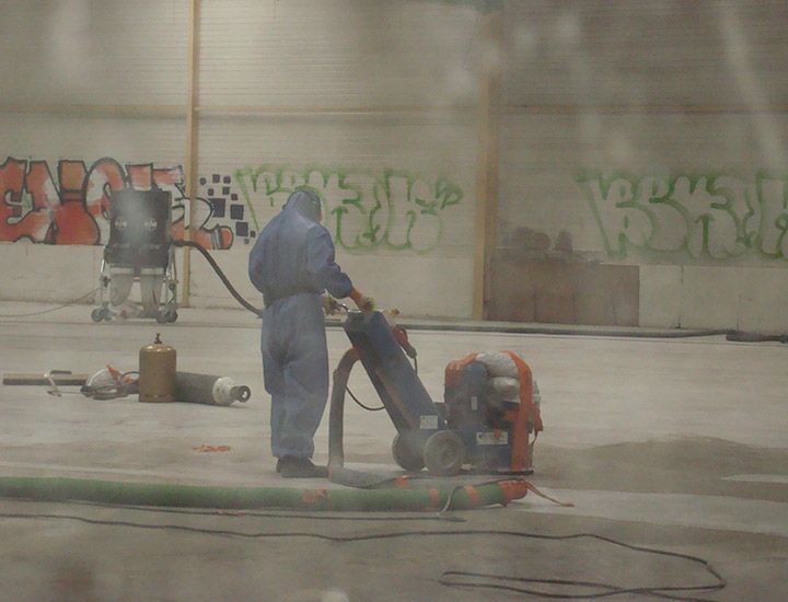 Une personne en combinaison de protection utilise une meuleuse à béton dans une grande pièce dont les murs sont recouverts de graffitis.