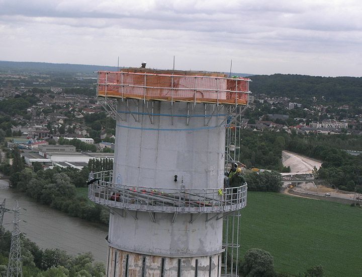 Construction d'une haute tour en béton avec échafaudages et barrières de sécurité orange, surplombant une rivière et une ville.