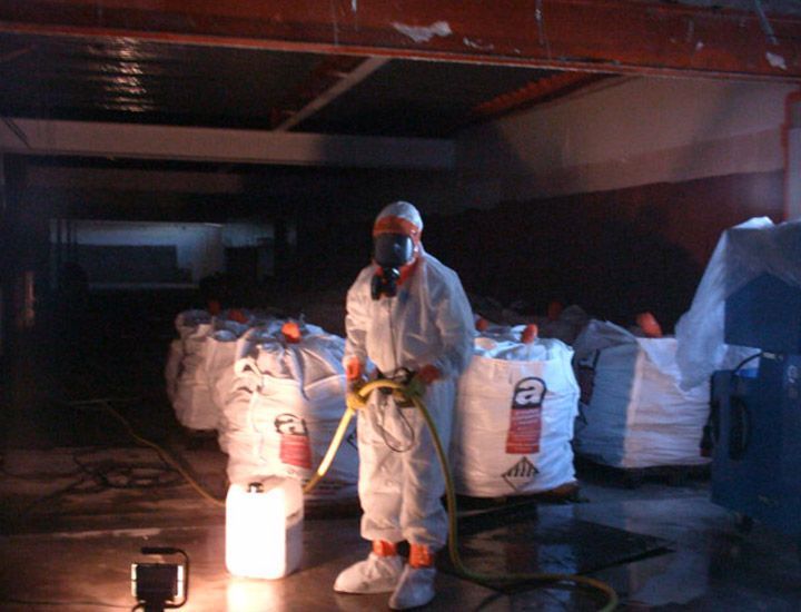 Une personne en combinaison de protection pulvérise de l'eau dans un entrepôt ; de grands sacs et conteneurs sont présents.