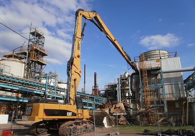 Une pelleteuse jaune démolit un bâtiment sur un site industriel par une journée ensoleillée.