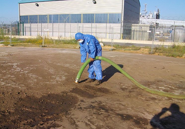 Personne en combinaison de protection bleue passant l'aspirateur sur de la terre sombre, en milieu industriel.
