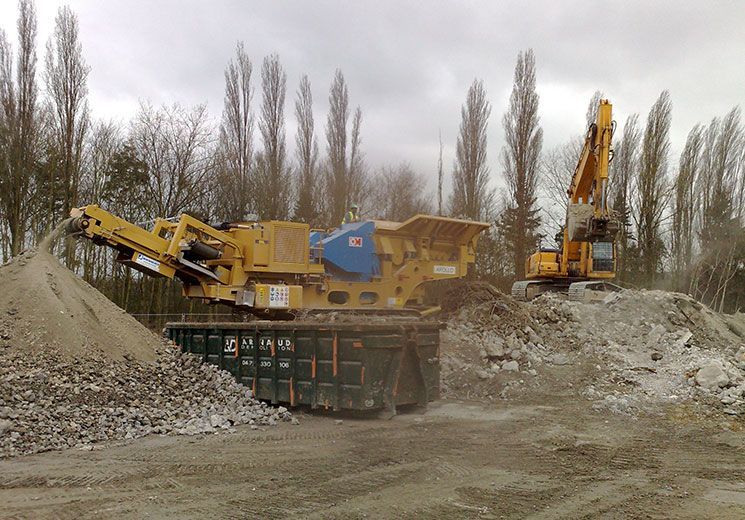 Concasseur de roches jaune traitant des débris, excavatrice sur un tas, en extérieur.