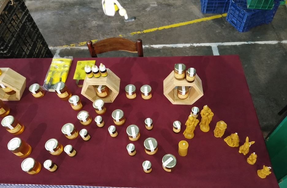 Productos de cera de abeja, como velas y tarros de miel, dispuestos sobre una mesa con un mantel granate en un evento.