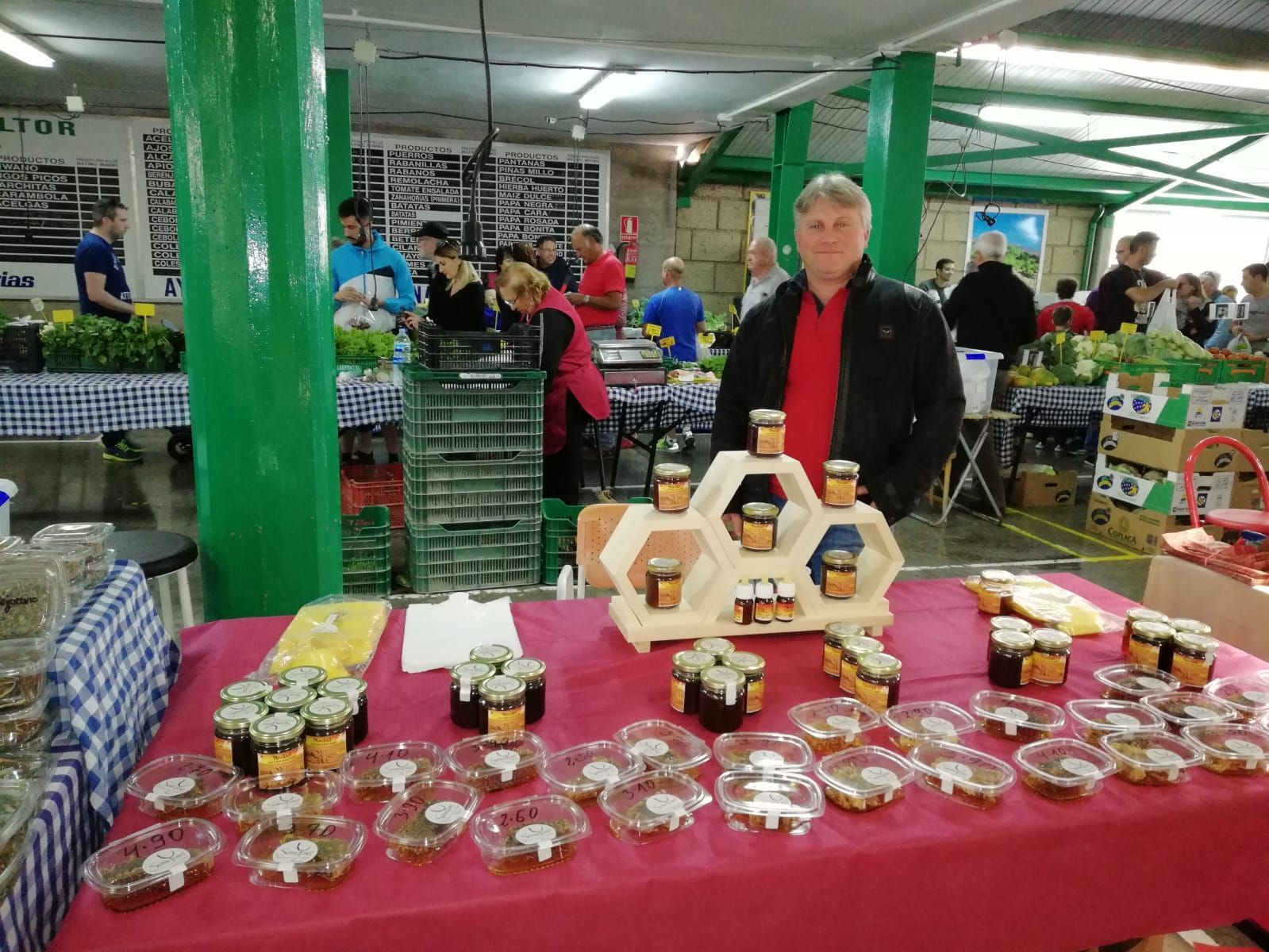 Una persona con camisa roja y chaqueta negra está de pie detrás de una mesa llena de frascos de miel en un mercado cubierto.
