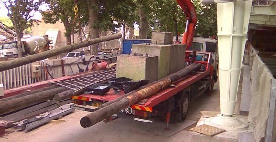 Camion utilitaire rouge stationné près d'un chantier, chargé de longs tuyaux métalliques et d'équipements.
